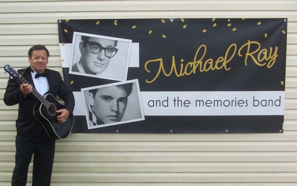 Man standing beside a banner for “Michael Ray and the memories band,” holding a guitar with two black-and-white portraits.